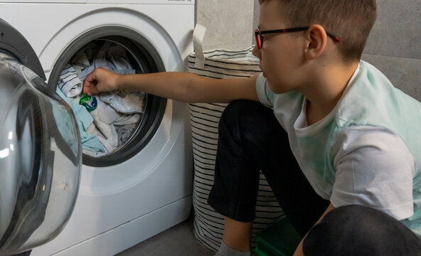 Boy with glasses putting laundry detergent pod capsule into washing machine drum with clothes in bathroom at home. Concept of children learning household tasks, independence skills and helping parents