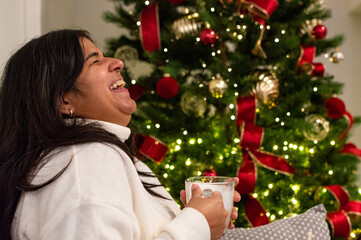 Smiling Woman Sitting by Decorated Christmas Tree. Woman Relaxing With Hot Drink by Christmas Tree.