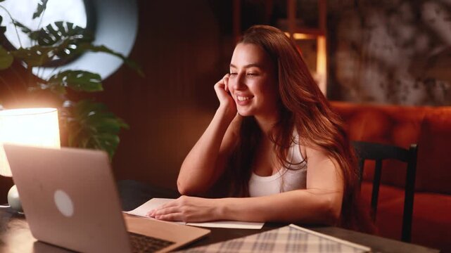 focused woman writing notes and smiling during late-night call, laptop and phone nearby, organized planner and pen, warm lamp light, productive remote work session, progress toward deadline