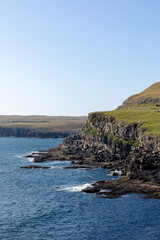 Naklejka premium Panoramic view of the dramatic coastline of the Faroe Islands, featuring steep volcanic cliffs, deep-blue North Atlantic waters and wide open green hills under a clear sky.