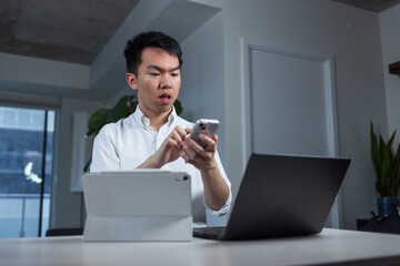 Young man juggles multiple devices while working multitasking from home. He focuses on his phone with a laptop and tablet on the desk.