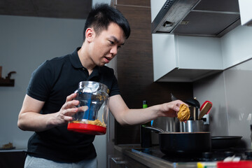 Young man prepares noodles on a stovetop at home, holding a container of dried pasta. His focused posture shows him adding ingredients to the boiling pot.