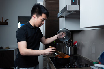Young man lifts a pot lid while stirring food on a stovetop in a home kitchen. The scene shows focused meal preparation with simple everyday tools.