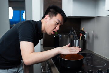 Young man leans over a hot pot and tests the rising heat with his hand in a home kitchen. The expression shows caution and focus during simple cooking.