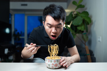 Young man leans over a bowl of noodles, lifting a portion with chopsticks preparing to eat dinner. Indoor home setting with a casual meal moment