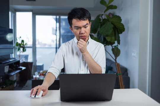 Young man in a white shirt concentrating while working on a laptop at home, hand on chin in deep thought. Represents remote work, productivity, and professional focus in a home office.