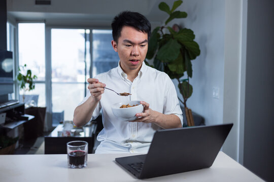 Young man eats a bowl of cereal at home while looking at his laptop. Shows a casual morning routine and remote work multitasking