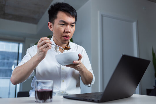 Young man eats cereal at home while focused on his laptop. Captures a casual morning routine and remote work multitasking. - Powered by Adobe