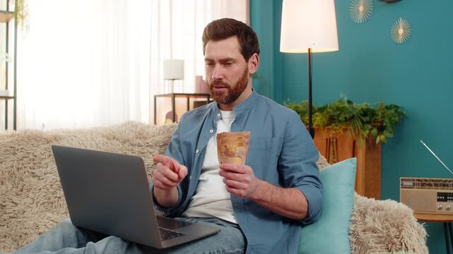 Young man at home holding money cash euros, looking thoughtfully at laptop screen while deciding on expenses. Caucasian guy sitting on sofa, considering purchases, investments, or financial planning
