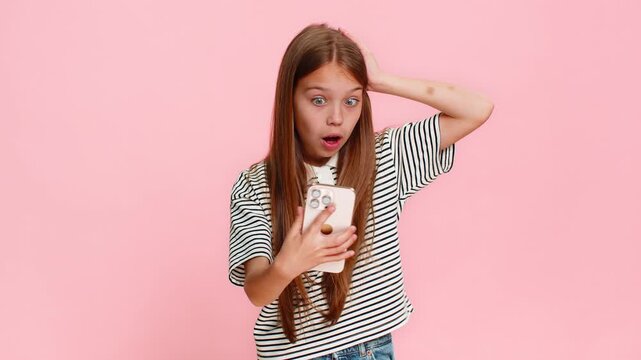 Young child girl reads a surprising message with unexpected news on smartphone, eyes wide open in astonishment clearly expressing disbelief shock, gasping with amazement. School kid on pink background