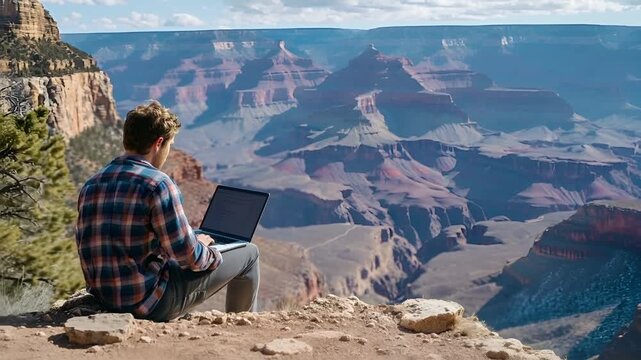 A man in a plaid shirt sits on a rocky outcrop overlooking a vast canyon, using a laptop. The sky above is partly cloudy, casting a soft light over the scene.