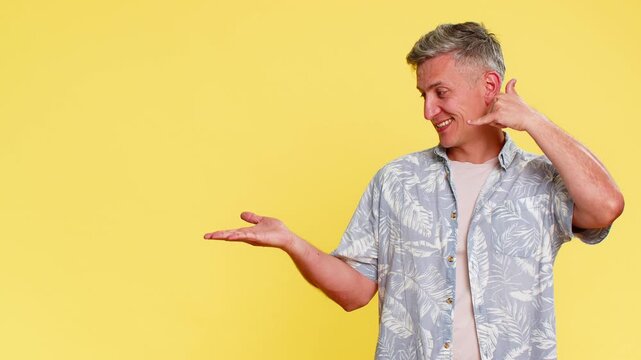 Middle-aged man making call me gesture near ear, then confidently pointing to left side, inviting to contact or call, clear intention. Guy expressing connection request isolated on yellow background.