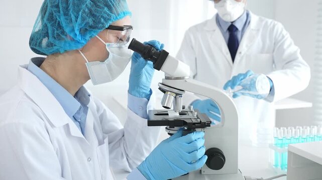 Female scientist wearing protective gear is examining specimen under microscope while man laboratory assistant handles glassware in a professional medical investigation. Medicine and science concept