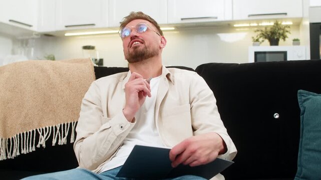 Young man at home drafts daily plan in notebook sets priorities and writes simple schedule for tasks. Caucasian guy on sofa lists goals adds time blocks and keeps focus with calm mood and clear mind.