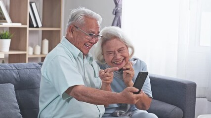 Asian elderly couple smiling and laughing on couch at home sharing smartphone video call and joyful health care moments in living room enjoying warm cozy connection