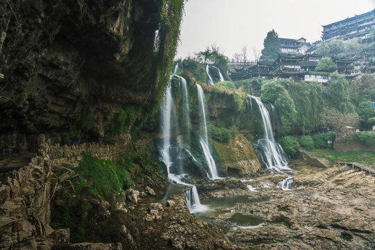 View of cascading waterfalls plunge over the rocky terrain, while ancient wooden buildings perch on the lush hillside, Xiangxi Tujia, Hunan, China.