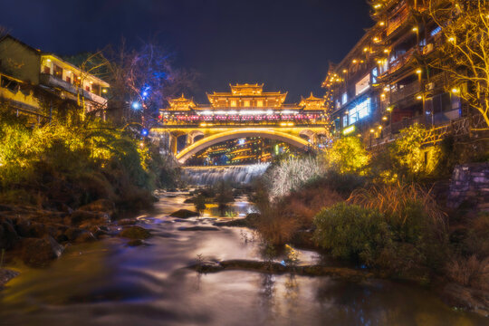 View of a bridge adorned with golden lights stretching over a flowing river, flanked by traditional buildings glowing warmly in the night, Xiangxi Tujia and Miao Autonomous Prefecture, Hunan, China.