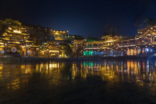 View of illuminated traditional buildings casting shimmering reflections on the dark water at night, Xiangxi Tujia and Miao Autonomous Prefecture, Hunan, China.