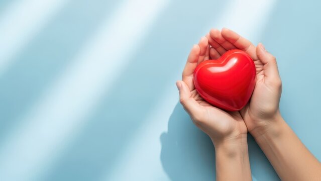 Pair of hands gently cupping and holding a shiny, bright red heart shape against a light blue background with diagonal shadow lines, symbolizing love, care, and health