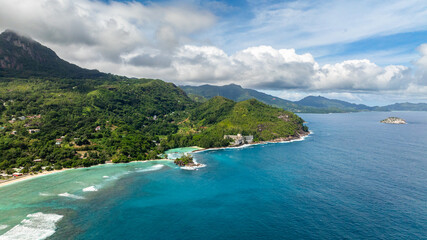 Coastal green hills with a turquoise ocean, small village structures, and sandy shorelines. Seychelles, Mahe.