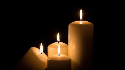 Four lit pillar candles glowing warmly in the dark, creating a solemn and peaceful atmosphere with soft light illuminating their wax surfaces against a deep black background
