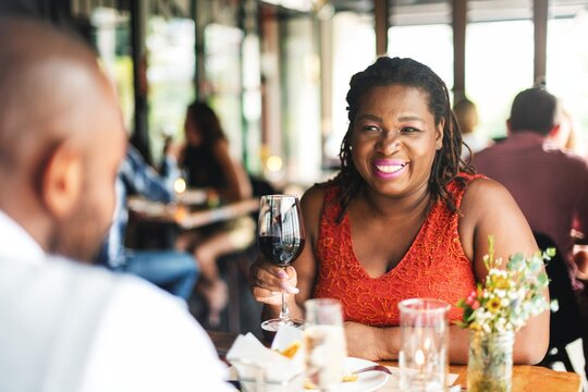 Smiling black woman enjoying wine at a restaurant. African American woman in a red dress, holding a wine glass. Dining, wine, and restaurant setting. Black couple celebrating at restaurant.