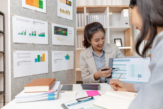 Two Asian businesswomen discussing ideas and analyzing reports from documents in a modern office. They collaborate on business strategies, teamwork, and organizational planning for success.
