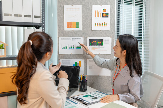 Two Asian businesswomen are smiling while discussing company performance reports in a modern office. They analyze data from printed documents, showing teamwork, planning, and collaboration in business