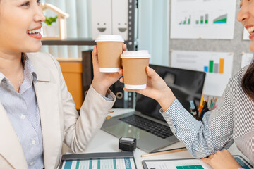 Two female colleagues take a short coffee break during their work meeting, smiling and clinking their coffee cups in a friendly gesture, showing teamwork, relaxation, and positive workplace culture.