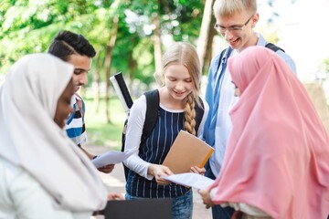 Obraz premium Group of diverse students outdoors, discussing papers. Smiling, engaged students in casual attire. Multicultural group enjoying study time together. International students and muslim girls at school.