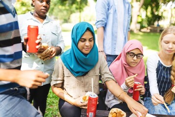 Group of diverse young adults enjoying a picnic outdoors. Diverse young women and young men sharing food and drinks, wearing casual attire, including hijabs, in a sunny park.