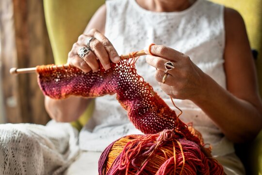 A senior woman knitting with orange yarn, focusing on hands and knitting needles. The knitting process is detailed, showing yarn texture and knitting technique. Senior woman knitting as hobby.