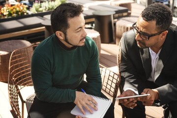 Two men in a business meeting outdoors, discussing ideas. One takes notes, the other holds a tablet. Business, meeting, discussion, ideas, outdoors. Men having conversation in cafe.