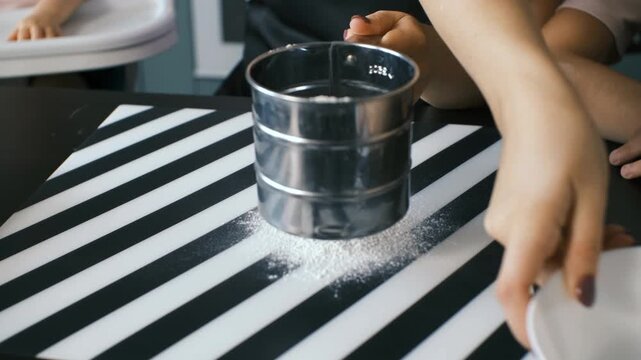 Fine white flour falling through a metal sifter onto a striped kitchen surface, creating a soft powdery layer as preparation for baking begins, baking ingredients and cozy home cooking concept