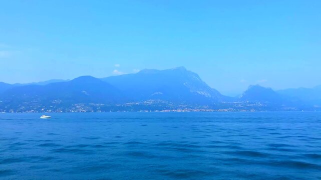 Mysterious scene from Lago di Garda with streaming deep blue waters, a white motorboat gently skimming them, and imposing mountainous peaks in the faraway distance, with human settlements underneath