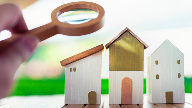 Hand holding magnifying glass over miniature wooden houses, symbolizing property search, house selection, real estate investment, home buying decisions, and market analysis concepts.