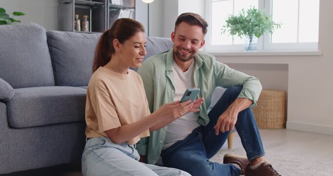 Couple using smartphone app at home. Seated together on the livingroom floor, they smile and browse the phone, showing relaxed planning in a bright interior. Concept digital lifestyle at home.