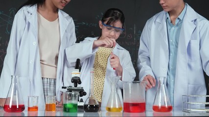 School girl mix colored solution while academic student support doing experiment at table with microscope and liquid. Diverse student standing at blackboard with science theory written. Edification - Powered by Adobe