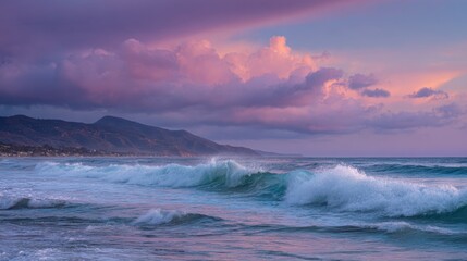 Fototapeta premium Colorful clouds reflect the setting sun over gentle ocean waves. Mountains rise in the distance completing the serene coastal scene during twilight hours.