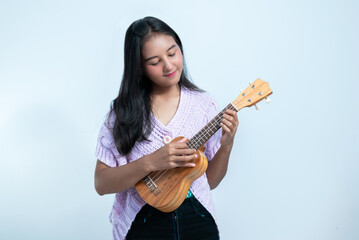 Fototapeta premium An Indonesian teenage girl holds a ukulele with a cheerful expression in a white studio background.