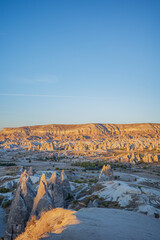 A striking vertical photograph of Cappadocia&rsquo;s iconic rock formations illuminated by warm sunrise light in Turkey. The golden cliffs, dramatic fairy chimneys, and expansive blue sky