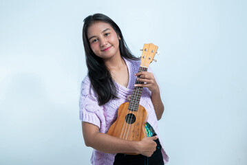 An Indonesian teenage girl holds a ukulele with a cheerful expression in a white studio background