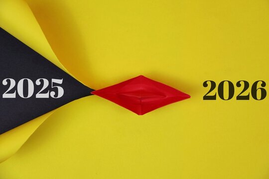 A yellow backdrop features a black folded shape meeting a red origami boat, representing the transition from 2025 to 2026. This image conveys renewal, progress, and optimistic planning for the future.