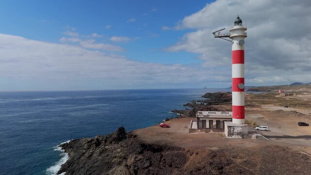 Aerial drone view flying past the iconic red and white striped Punta de Abona Lighthouse on the arid coast of Tenerife, Canary Islands, Spain.