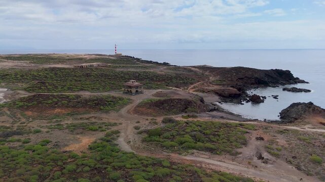 Wide aerial drone shot showing the Punta de Abona Lighthouse standing near the desolate ruins of the abandoned Abades Leper Colony on the arid coast of Tenerife, Spain.
