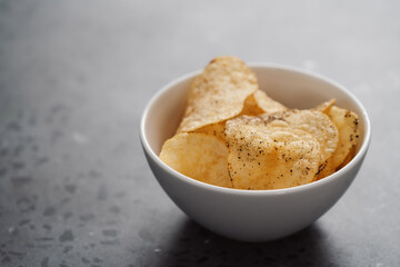 White bowl with organic potato chips sprinkled with fresh ground black pepper on concrete background with copy space