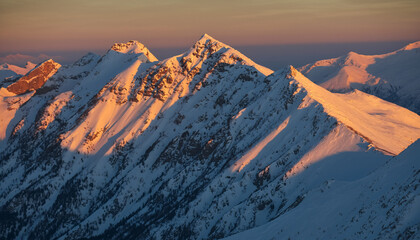 Snow-capped mountain peaks bathed in the warm glow of sunrise or sunset, creating a dramatic alpine landscape.