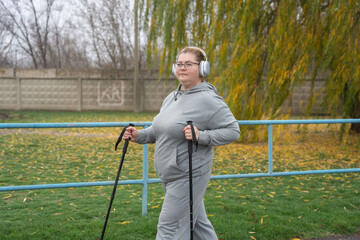 Woman in a grey tracksuit and headphones practicing nordic walking with poles exercising outdoors in a determined weight-loss program