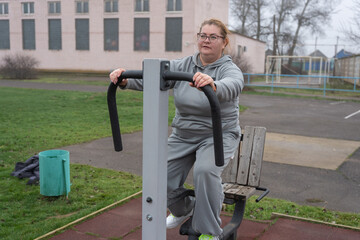 An overweight woman exercises seated on an outdoor exercise machine, seeking to lose weight, strengthen muscles, improve her health, and boost her confidence