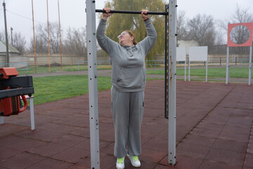 Plus-size woman doing pull-up on outdoor bar in gray hoodie and sweatpants, showing perseverance and self-care while building strength and balance during a cool morning in a public park gym